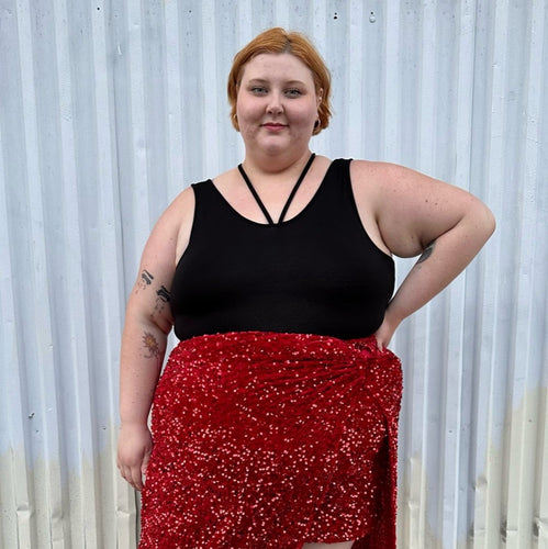 Front view of a size 2 Torrid black criss-cross detail stretchy bodysuit styled tucked into a red sequin maxi skirt on a size 22/24 model. The photo is taken outside in natural lighting.