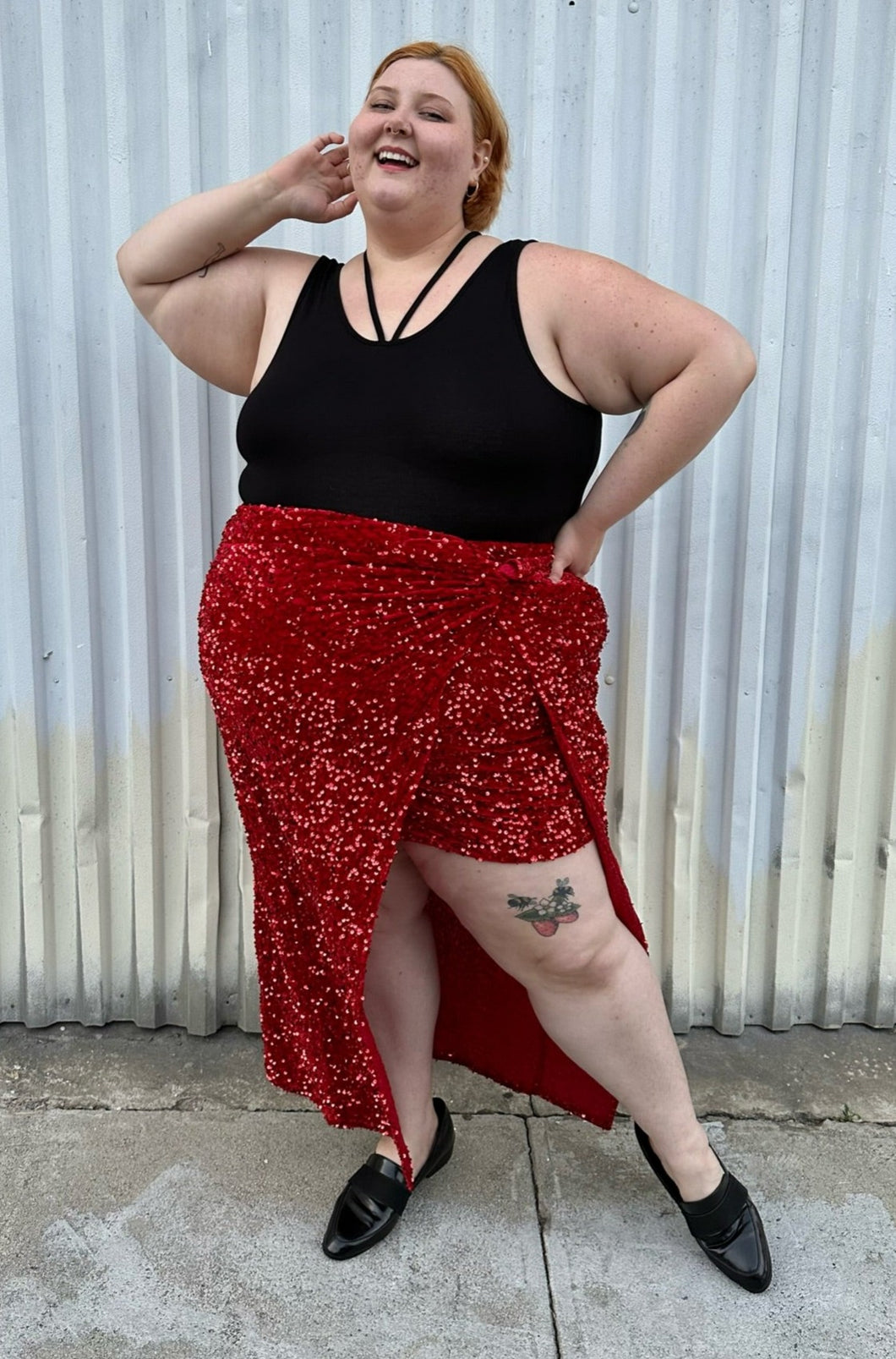 Full-body front view of a size 24 Eloquii red sequin twist-detail maxi skirt with high open slit and mini slip skirt underneath styled with a black criss-cross top and black loafers on a size 22/24 model. The photo is taken outside in natural lighting.