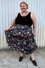 Load image into Gallery viewer, Full-body front view of a size 24 Eloquii black pleather maxi skirt with cool-toned floral all-over pattern styled with a black criss-cross tank tucked in and black loafers on a size 22/24 model. The photo is taken outside in natural lighitng.
