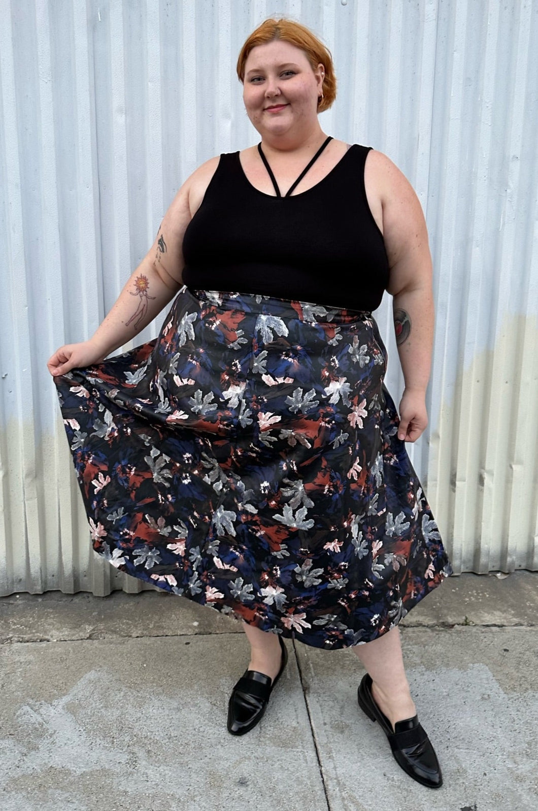Full-body front view of a size 24 Eloquii black pleather maxi skirt with cool-toned floral all-over pattern styled with a black criss-cross tank tucked in and black loafers on a size 22/24 model. The photo is taken outside in natural lighitng.