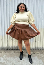 Load image into Gallery viewer, Full-body front view of a size 20 Eloquii brown pleather pleated mini skirt styled with a cream turtleneck sweater on a size 18/20 model. The photo is taken outside in natural lighting.
