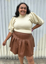 Load image into Gallery viewer, Front view of a size 20 Eloquii brown pleather pleated mini skirt styled with a cream turtleneck sweater on a size 18/20 model. The photo is taken outside in natural lighting.
