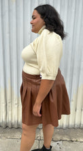 Load image into Gallery viewer, Side view of a size 20 Eloquii brown pleather pleated mini skirt styled with a cream turtleneck sweater on a size 18/20 model. The photo is taken outside in natural lighting.
