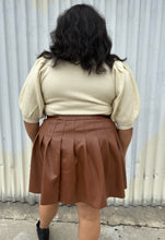 Load image into Gallery viewer, Back view of a size 20 Eloquii brown pleather pleated mini skirt styled with a cream turtleneck sweater on a size 18/20 model. The photo is taken outside in natural lighting.
