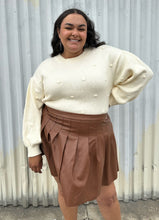 Load image into Gallery viewer, Additional front view of a size 20 Eloquii brown pleather pleated mini skirt styled with a cream turtleneck sweater on a size 18/20 model. The photo is taken outside in natural lighting.
