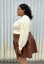 Load image into Gallery viewer, Side view of a size 18/20 Eloquii cream chunky knit sweater with swiss dots styled tucked into a brown pleather mini skirt on a size 18/20 model. The photo is taken outside in natural lighting.
