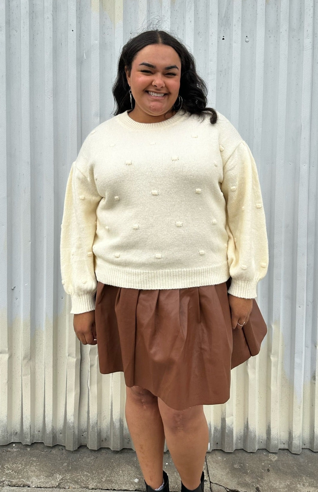 Front view of a size 18/20 Eloquii cream chunky knit sweater with swiss dots styled over a brown pleather mini skirt on a size 18/20 model. The photo is taken outside in natural lighting.