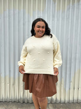Load image into Gallery viewer, Additional front view of a size 18/20 Eloquii cream chunky knit sweater with swiss dots styled over a brown pleather mini skirt on a size 18/20 model. The photo is taken outside in natural lighting.
