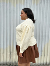 Load image into Gallery viewer, Side view of a size 18/20 Eloquii cream chunky knit sweater with swiss dots styled over a brown pleather mini skirt on a size 18/20 model. The photo is taken outside in natural lighting.

