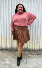 Load image into Gallery viewer, Full-body front view of a size 18/20 Eloquii blush pink cable-knit turtleneck sweater with thick fold-over neck and swiss dots styled tucked into a brown pleather mini skirt and black boots on a size 18/20 model. The photo is taken outside in natural lighting.
