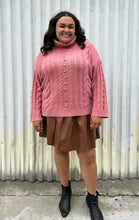 Load image into Gallery viewer, Full-body front view of a size 18/20 Eloquii blush pink cable-knit turtleneck sweater with thick fold-over neck and swiss dots styled over a brown pleather mini skirt and black boots on a size 18/20 model. The photo is taken outside in natural lighting.
