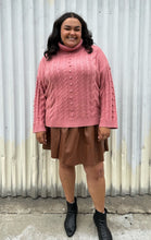 Load image into Gallery viewer, Full-body front view of a size 18/20 Eloquii blush pink cable-knit turtleneck sweater with thick fold-over neck and swiss dots styled over a brown pleather mini skirt and black boots on a size 18/20 model. The photo is taken outside in natural lighting.
