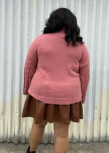 Load image into Gallery viewer, Back view of a size 18/20 Eloquii blush pink cable-knit turtleneck sweater with thick fold-over neck and swiss dots styled over a brown pleather mini skirt on a size 18/20 model. The photo is taken outside in natural lighting.
