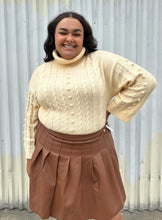 Load image into Gallery viewer, Front view of a size 18/20 Eloquii cream cable-knit turtleneck sweater with thick fold-over neck and swiss dots styled tucked into a brown pleather mini skirt on a size 18/20 model. The photo is taken outside in natural lighting.
