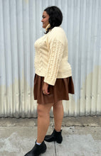 Load image into Gallery viewer, Full-body side view of a size 18/20 Eloquii cream cable-knit turtleneck sweater with thick fold-over neck and swiss dots styled over a brown pleather mini skirt and black boots on a size 18/20 model. The photo is taken outside in natural lighting.
