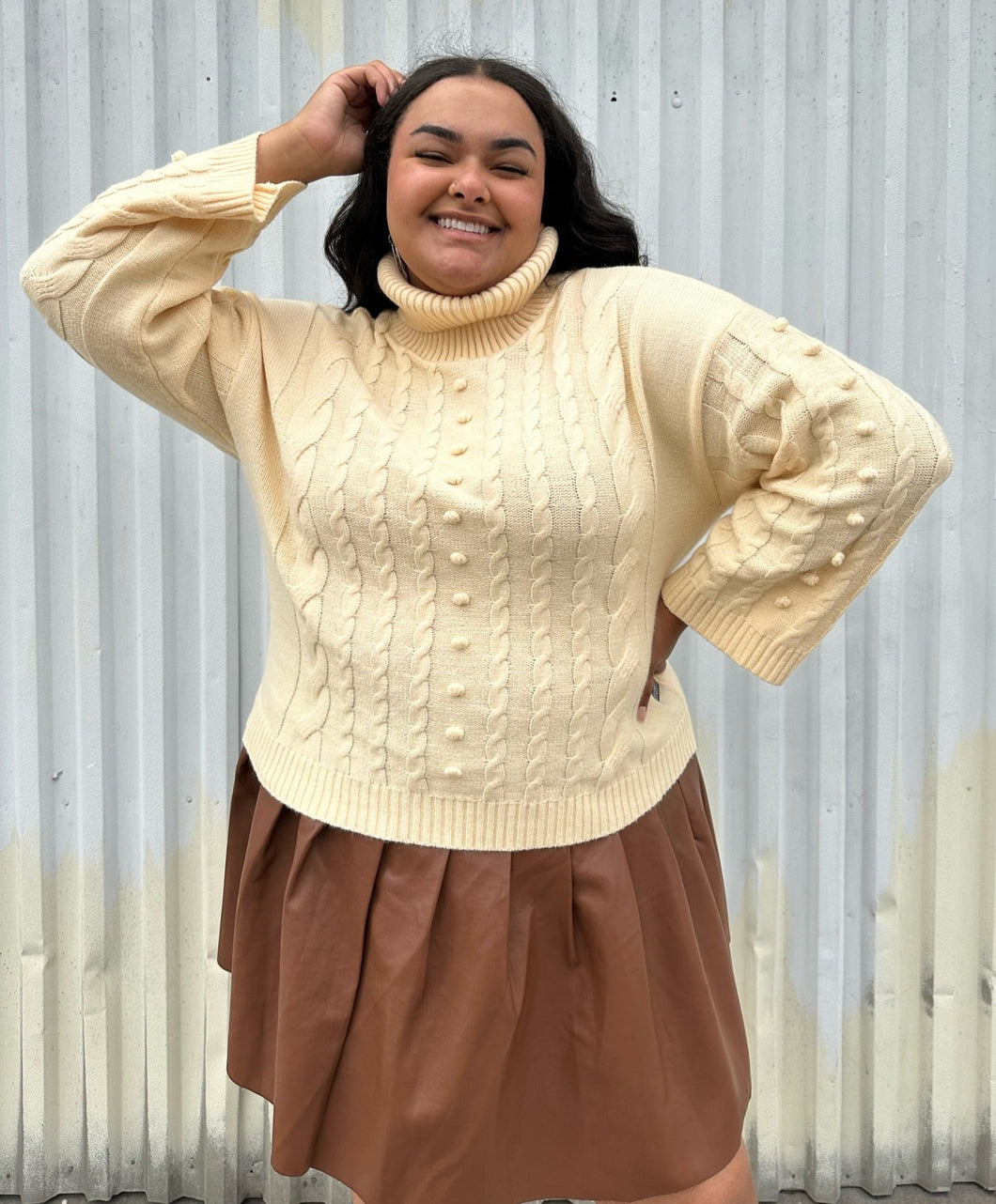 Front view of a size 18/20 Eloquii cream cable-knit turtleneck sweater with thick fold-over neck and swiss dots styled over a brown pleather mini skirt on a size 18/20 model. The photo is taken outside in natural lighting.