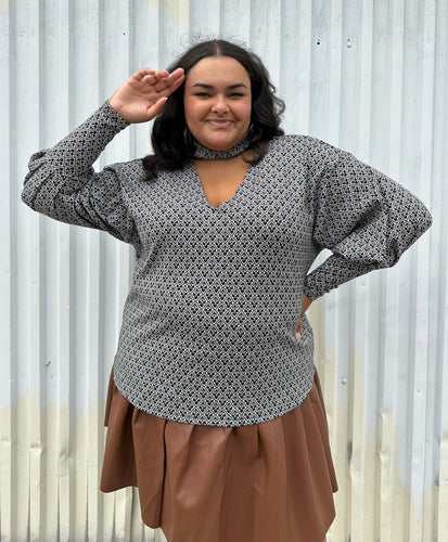 Front view of a size 20 Eloquii black and white patterned sweater with choker neckline and puff sleeves styled over a brown pleather mini skirt on a size 18/20 model. The photo is taken outside in natural lighting.