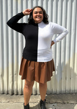 Load image into Gallery viewer, Full-body view of a size 18/20 Eloquii black and white halfsies long sleeve tee styled over a brown pleather mini skirt with black boots on a size 18/20 model. The photo is taken outside in natural lighting.
