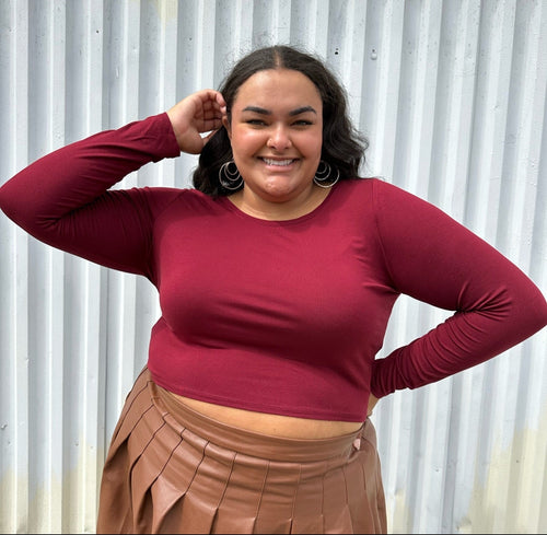 Front view of a size 18/20 maroon long sleeve cropped t-shirt styled over a brown pleather mini skirt on a size 18/20 model. The photo is taken outside in natural lighting.