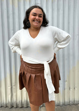 Load image into Gallery viewer, Front view of a size 18/20 Eloquii off-white longline sweater with side slits, a tie belt, subtle puff sleeves, and a v neck styled tucked into a brown pleather mini skirt on a size 18/20 model. The photo is taken outside in natural lighting.
