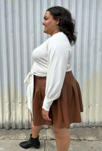 Load image into Gallery viewer, Side view of a size 18/20 Eloquii off-white longline sweater with side slits, a tie belt, subtle puff sleeves, and a v neck styled tucked into a brown pleather mini skirt on a size 18/20 model. The photo is taken outside in natural lighting.
