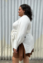Load image into Gallery viewer, Side view of a size 18/20 Eloquii off-white longline sweater with side slits, a tie belt, subtle puff sleeves, and a v neck styled over a brown pleather mini skirt on a size 18/20 model. The photo is taken outside in natural lighting.
