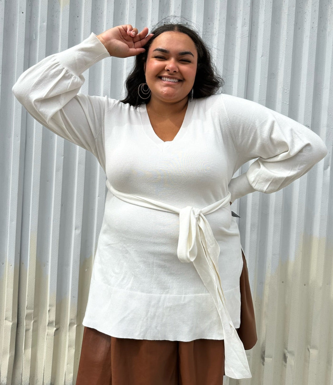 Front view of a size 18/20 Eloquii off-white longline sweater with side slits, a tie belt, subtle puff sleeves, and a v neck styled over a brown pleather mini skirt on a size 18/20 model. The photo is taken outside in natural lighting.