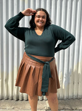 Load image into Gallery viewer, Front view of a size 18/20 Eloquii dark green longline sweater with side slits, a tie belt, subtle puff sleeves, and a v neck styled tucked into a brown pleather mini skirt on a size 18/20 model. The photo is taken outside in natural lighting.

