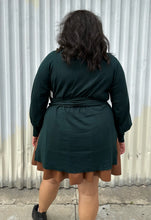 Load image into Gallery viewer, Back view of a size 18/20 Eloquii dark green longline sweater with side slits, a tie belt, subtle puff sleeves, and a v neck styled over a brown pleather mini skirt on a size 18/20 model. The photo is taken outside in natural lighting.
