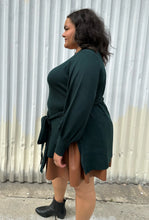 Load image into Gallery viewer, Side view of a size 18/20 Eloquii dark green longline sweater with side slits, a tie belt, subtle puff sleeves, and a v neck styled over a brown pleather mini skirt on a size 18/20 model. The photo is taken outside in natural lighting.
