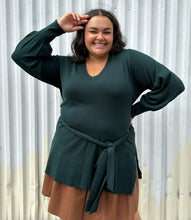 Load image into Gallery viewer, Front view of a size 18/20 Eloquii dark green longline sweater with side slits, a tie belt, subtle puff sleeves, and a v neck styled over a brown pleather mini skirt on a size 18/20 model. The photo is taken outside in natural lighting.
