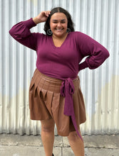 Load image into Gallery viewer, Front view of a size 18/20 Eloquii berry pink longline sweater with side slits, a tie belt, subtle puff sleeves, and a v neck styled tucked into a brown pleather mini skirt on a size 18/20 model. The photo is taken outside in natural lighting.
