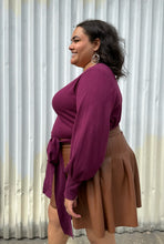 Load image into Gallery viewer, Side view of a size 18/20 Eloquii berry pink longline sweater with side slits, a tie belt, subtle puff sleeves, and a v neck styled tucked into a brown pleather mini skirt on a size 18/20 model. The photo is taken outside in natural lighting.
