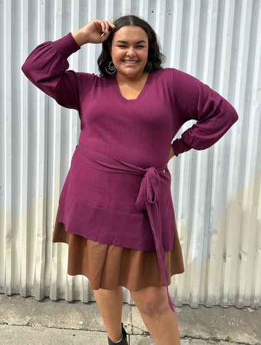 Front view of a size 18/20 Eloquii berry pink longline sweater with side slits, a tie belt, subtle puff sleeves, and a v neck styled over a brown pleather mini skirt on a size 18/20 model. The photo is taken outside in natural lighting.