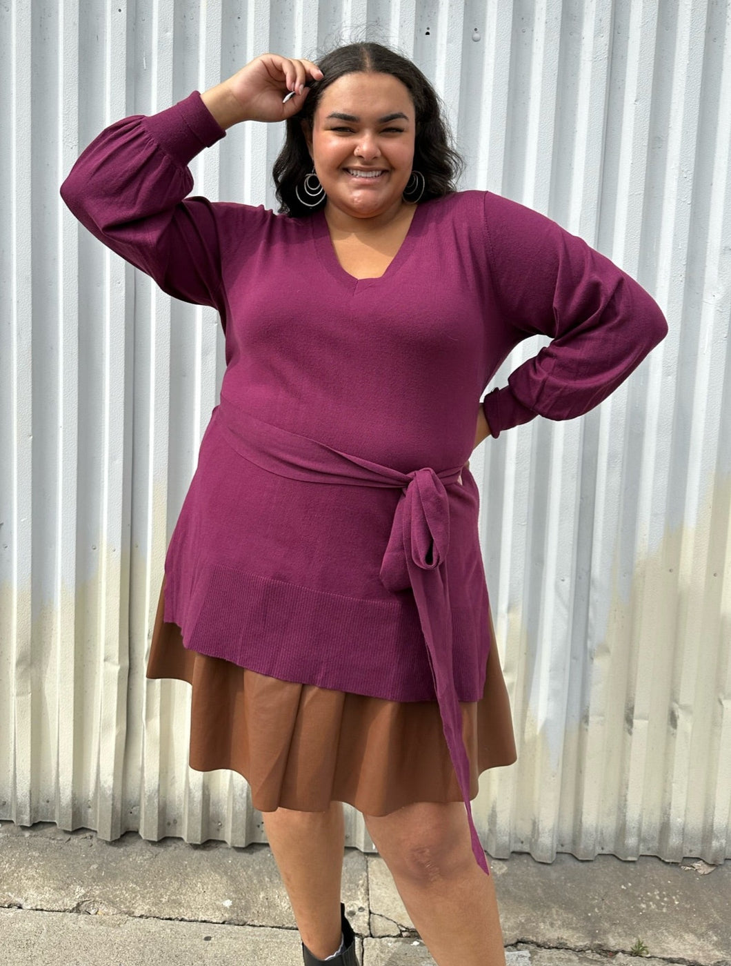 Front view of a size 18/20 Eloquii berry pink longline sweater with side slits, a tie belt, subtle puff sleeves, and a v neck styled over a brown pleather mini skirt on a size 18/20 model. The photo is taken outside in natural lighting.