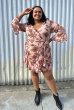 Load image into Gallery viewer, Additional full-body front view of a size 18 City Chic pale pink wrap dress with red and green florals, lace details, and bell sleeves styled with black boots on a size 18/20 model. The photo is taken outside in natural lighting.
