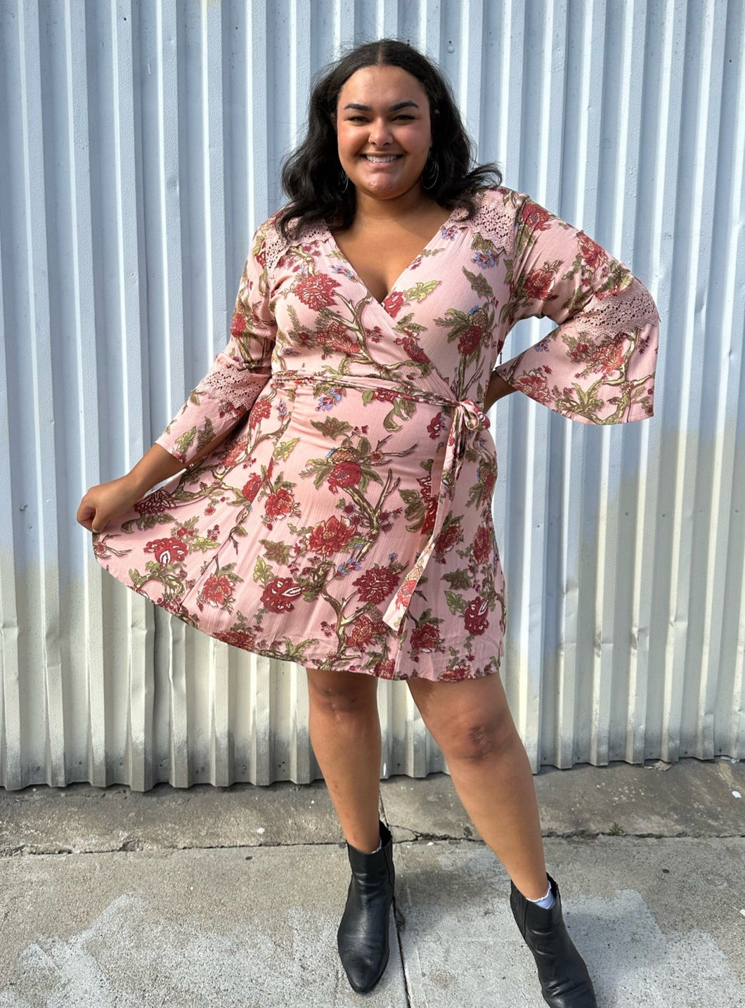 Full-body front view of a size 18 City Chic pale pink wrap dress with red and green florals, lace details, and bell sleeves styled with black boots on a size 18/20 model. The photo is taken outside in natural lighting.