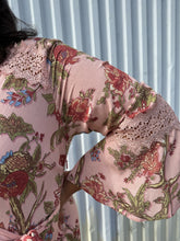 Load image into Gallery viewer, Close up view of the lace detail and bell sleeves of a size 18 City Chic pale pink wrap dress with red and green florals, lace details, and bell sleeves on a size 18/20 model. The photo is taken outside in natural lighting.
