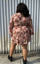 Load image into Gallery viewer, Full-body back view of a size 18 City Chic pale pink wrap dress with red and green florals, lace details, and bell sleeves styled with black boots on a size 18/20 model. The photo is taken outside in natural lighting.
