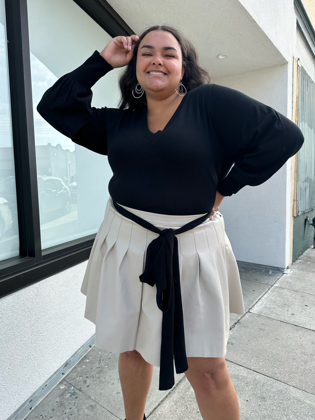 Front view of a size 18/20 Eloquii black longline sweater with side slits, a tie belt, subtle puff sleeves, and a v neck styled tucked into a brown pleather mini skirt on a size 18/20 model. The photo is taken outside in natural lighting.