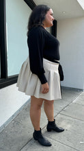 Load image into Gallery viewer, Full-body side view of a size 20 Eloquii off-white pleather pleated mini skirt styled with a black sweater on a size 18/20 model. The photo is taken outside in natural lighting.
