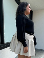 Load image into Gallery viewer, Side view of a size 18/20 Eloquii black longline sweater with side slits, a tie belt, subtle puff sleeves, and a v neck styled tucked into a brown pleather mini skirt on a size 18/20 model. The photo is taken outside in natural lighting.
