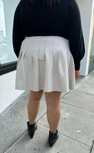 Load image into Gallery viewer, Back view of a size 20 Eloquii off-white pleather pleated mini skirt styled with a black sweater on a size 18/20 model. The photo is taken outside in natural lighting.
