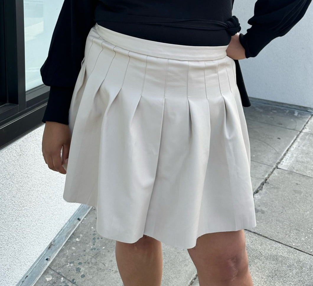 Front view of a size 20 Eloquii off-white pleather pleated mini skirt styled with a black sweater on a size 18/20 model. The photo is taken outside in natural lighting.
