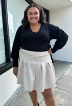 Load image into Gallery viewer, Front view of a size 20 Eloquii off-white pleather pleated mini skirt styled with a black sweater on a size 18/20 model. The photo is taken outside in natural lighting.
