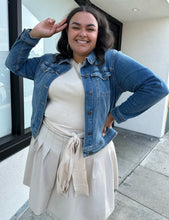 Load image into Gallery viewer, Front view of a size 1X Ava &amp; Viv medium wash denim jacket styled open over a cream sweater and mini skirt on a size 18/20 model. The photo is taken outside in natural lighting.
