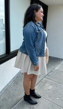 Load image into Gallery viewer, Full-body side view of a size 1X Ava &amp; Viv medium wash denim jacket styled open over a cream sweater and mini skirt on a size 18/20 model. The photo is taken outside in natural lighting.
