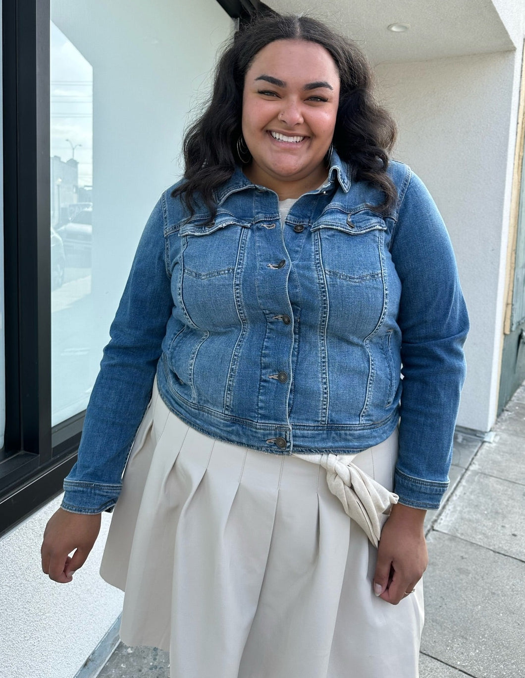Front view of a size 1X Ava & Viv medium wash denim jacket styled buttoned up over a cream sweater and mini skirt on a size 18/20 model. The photo is taken outside in natural lighting.