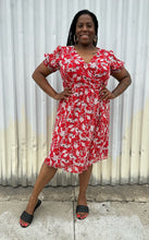Load image into Gallery viewer, Additional full-body front view of a size L Chelsea28 red, white, and black floral wrap midi dress with subtle ruffles and puff at the shoulders styled with black slides on a size 14/16 model. The photo is taken outside in natural lighting.
