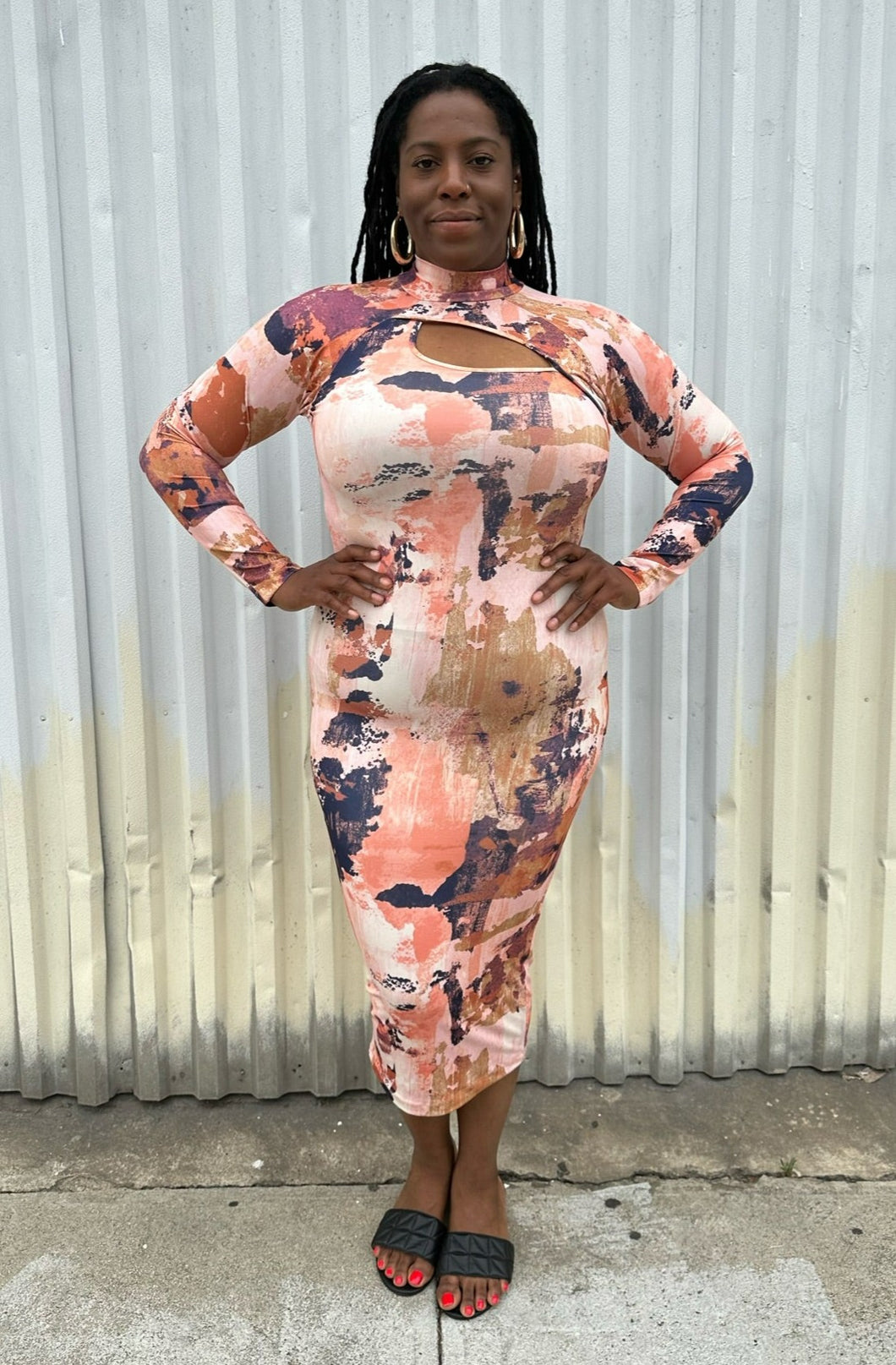 Full-body front view of a size 0X Nina Parker peach, orange, cream, and purple abstract pattern asymmetrical cut-out bust long sleeve bodycon midi dress styled with black slides on a size 14/16 model. The photo is taken outside in natural lighting.